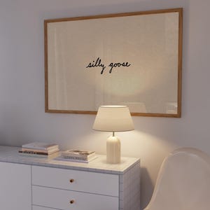 May include: A framed artwork with the words "silly goose" in cursive script hangs above a white cabinet. A white lamp with a lampshade sits on the cabinet, next to a stack of books. A white chair is in the foreground.