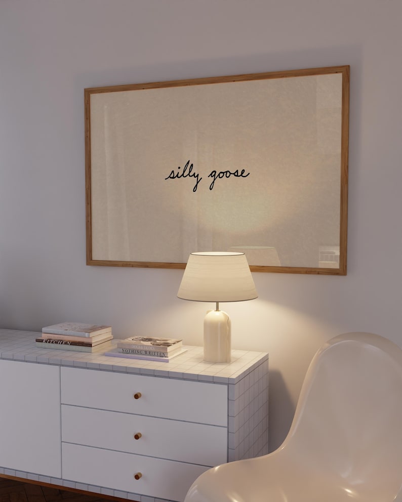 May include: A framed artwork with the words "silly goose" in cursive script hangs above a white cabinet. A white lamp with a lampshade sits on the cabinet, next to a stack of books. A white chair is in the foreground.