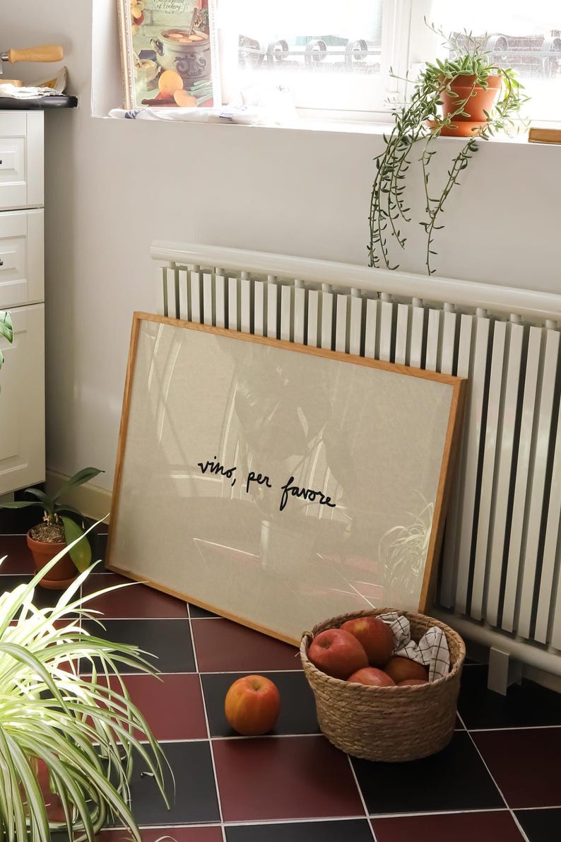 May include: A framed artwork with the text "vino, per favore" sits against a white radiator. A woven basket filled with red apples and a single apple are on a black and red tiled floor. A potted plant is in the foreground.