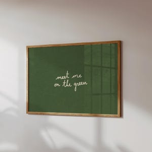 May include: A framed art print with a green background and the handwritten text "meet me on the green" in white. The frame is made of wood. The artwork is mounted on a white wall.