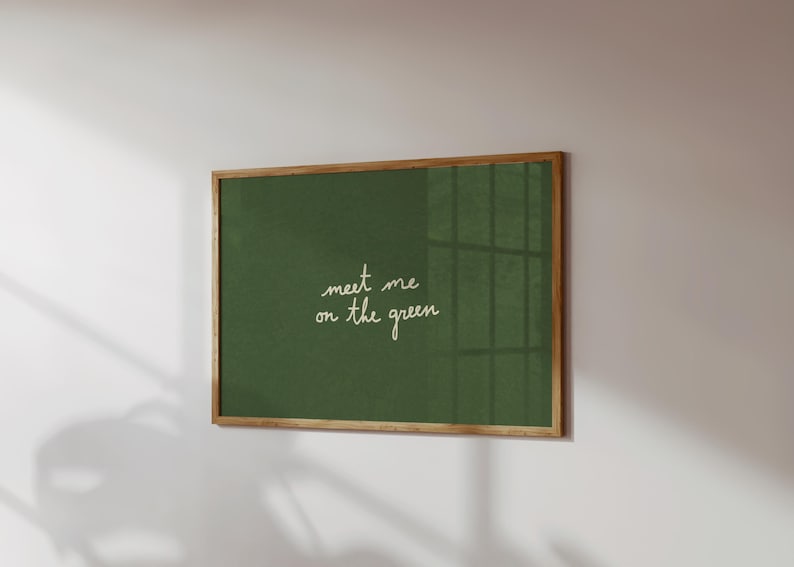 May include: A framed art print with a green background and the handwritten text "meet me on the green" in white. The frame is made of wood. The artwork is mounted on a white wall.