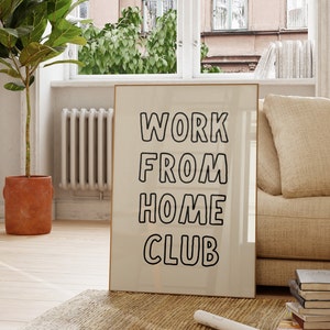Op de afbeelding: Een ingelijste print met de woorden "WORK FROM HOME CLUB" in zwarte, vette letters. De print leunt tegen een muur op een jute vloerkleed, in de buurt van een raam met uitzicht op een gebouw. Een potplant en een bank staan ook in de kamer.