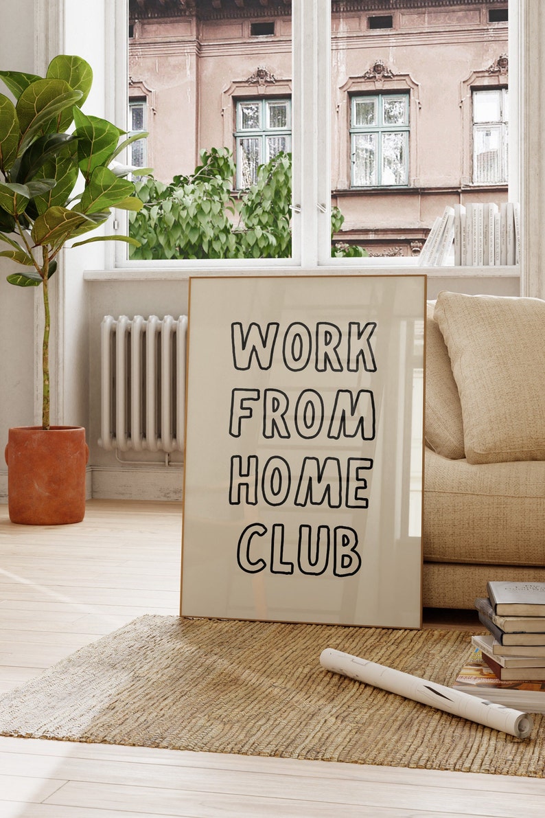 Op de afbeelding: Een ingelijste print met de woorden "WORK FROM HOME CLUB" in zwarte, vette letters. De print leunt tegen een muur op een jute vloerkleed, in de buurt van een raam met uitzicht op een gebouw. Een potplant en een bank staan ook in de kamer.