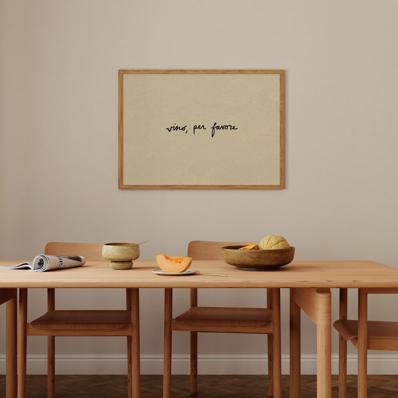 May include: A dining room scene featuring a wooden table and chairs. A framed print on the wall reads "vino, per favore" in black script. The table is set with a bowl, a plate with melon, and a rolled-up newspaper.