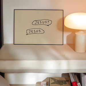 May include: A framed artwork with the text "JESUS?" and "JESUS." in speech bubbles. The artwork is displayed on a white surface, next to a white table lamp with a rounded shade and a red book.