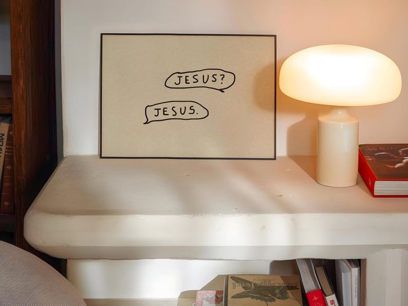 May include: A framed artwork with the text "JESUS?" and "JESUS." in speech bubbles. The artwork is displayed on a white surface, next to a white table lamp with a rounded shade and a red book.