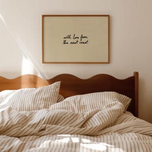 Puede incluir: Una impresi&oacute;n enmarcada con el texto "with love from the east coast" cuelga sobre una cama con ropa de cama a rayas. El marco es de color marr&oacute;n c&aacute;lido y la ropa de cama tiene una paleta de colores neutros.