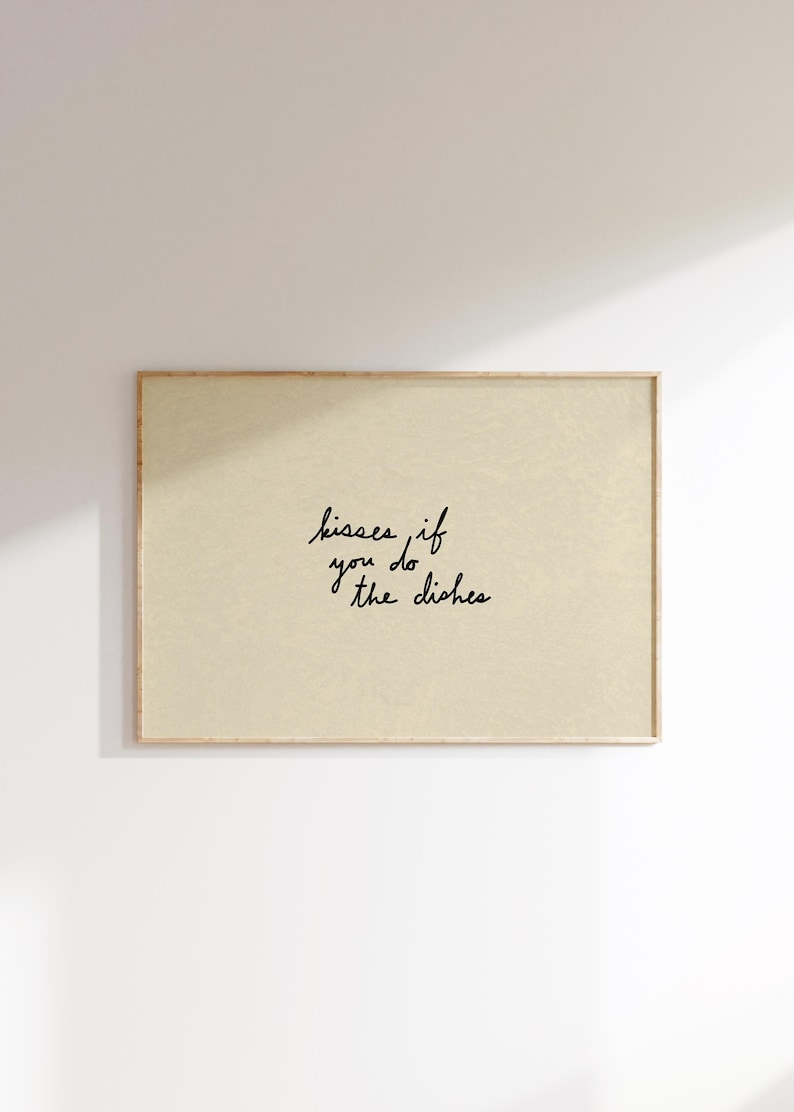 May include: A framed print with a light beige background and the handwritten text "kisses if you do the dishes" in black cursive. The frame is a light brown color.