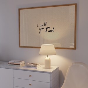 Puede incluir: Una obra de arte enmarcada con el texto "i will give you rest" cuelga sobre un mueble blanco. Una l&aacute;mpara blanca con una pantalla beige est&aacute; sobre el mueble, junto a una pila de libros. Una silla blanca est&aacute; en primer plano.