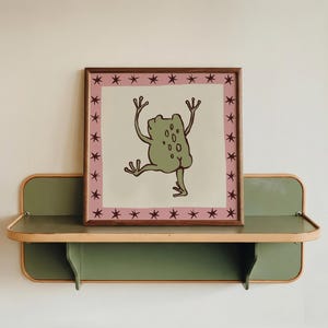 May include: A square framed print featuring a light green frog illustration with raised arms and legs on a cream background. The border is a dusty pink square decorated with small dark brown stars, all enclosed in a simple wooden frame.