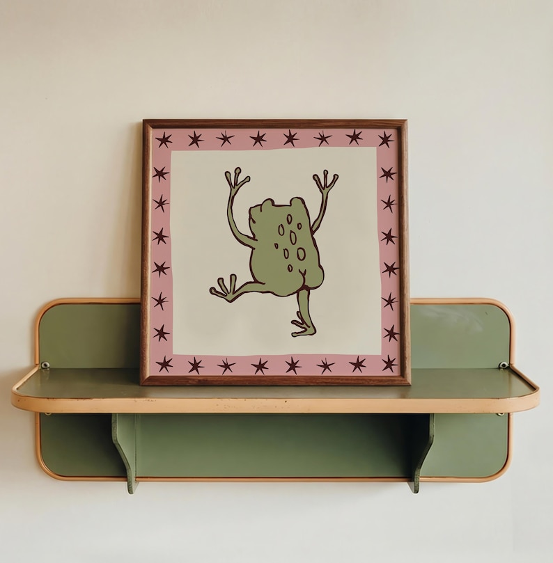 May include: A square framed print featuring a light green frog illustration with raised arms and legs on a cream background. The border is a dusty pink square decorated with small dark brown stars, all enclosed in a simple wooden frame.