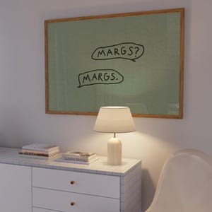 May include: A framed artwork with the text "MARGS?" and "MARGS." in black on a green background. A white lamp with a beige lampshade sits on a white cabinet with gold knobs. Books are stacked on the cabinet. A white chair is in the foreground.