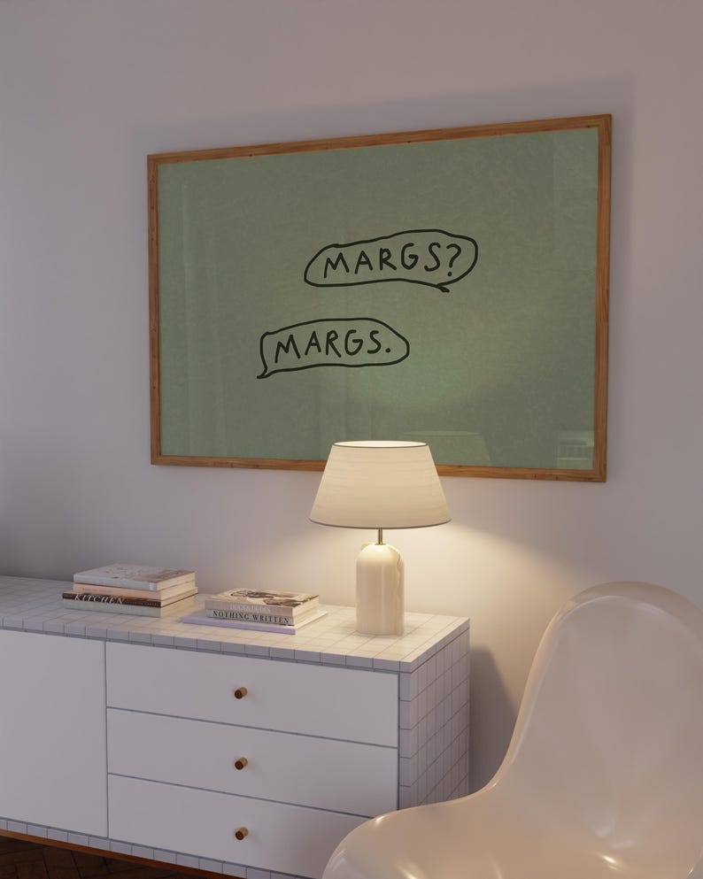 May include: A framed artwork with the text "MARGS?" and "MARGS." in black on a green background. A white lamp with a beige lampshade sits on a white cabinet with gold knobs. Books are stacked on the cabinet. A white chair is in the foreground.