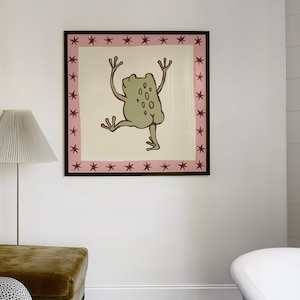 May include: A square framed wall art print featuring a light green frog illustration centred on a cream background. The artwork is enclosed by a pink border decorated with a repeating pattern of dark maroon stars, all set within a thin black frame.