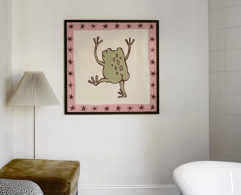 May include: A square framed wall art print featuring a light green frog illustration centred on a cream background. The artwork is enclosed by a pink border decorated with a repeating pattern of dark maroon stars, all set within a thin black frame.