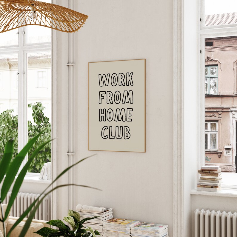 Op de afbeelding: Een ingelijste poster met de tekst "WORK FROM HOME CLUB" in zwarte, omlijnde letters. De poster is beige en hangt aan een witte muur. De kamer heeft grote ramen, planten en stapels tijdschriften.