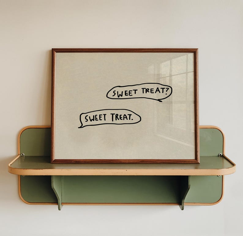 May include: A framed print with the text "SWEET TREAT?" and "SWEET TREAT." in black speech bubbles. The frame is brown, and the print is displayed on a green and tan shelf against a white wall.