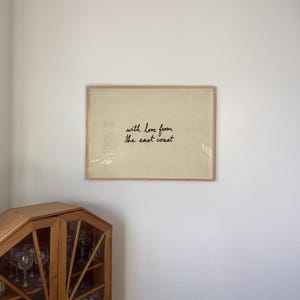Puede incluir: Una impresi&oacute;n enmarcada con el texto "with love from the east coast" en escritura negra. La impresi&oacute;n tiene un fondo beige claro y un marco de madera natural. La impresi&oacute;n est&aacute; colgada en una pared blanca.