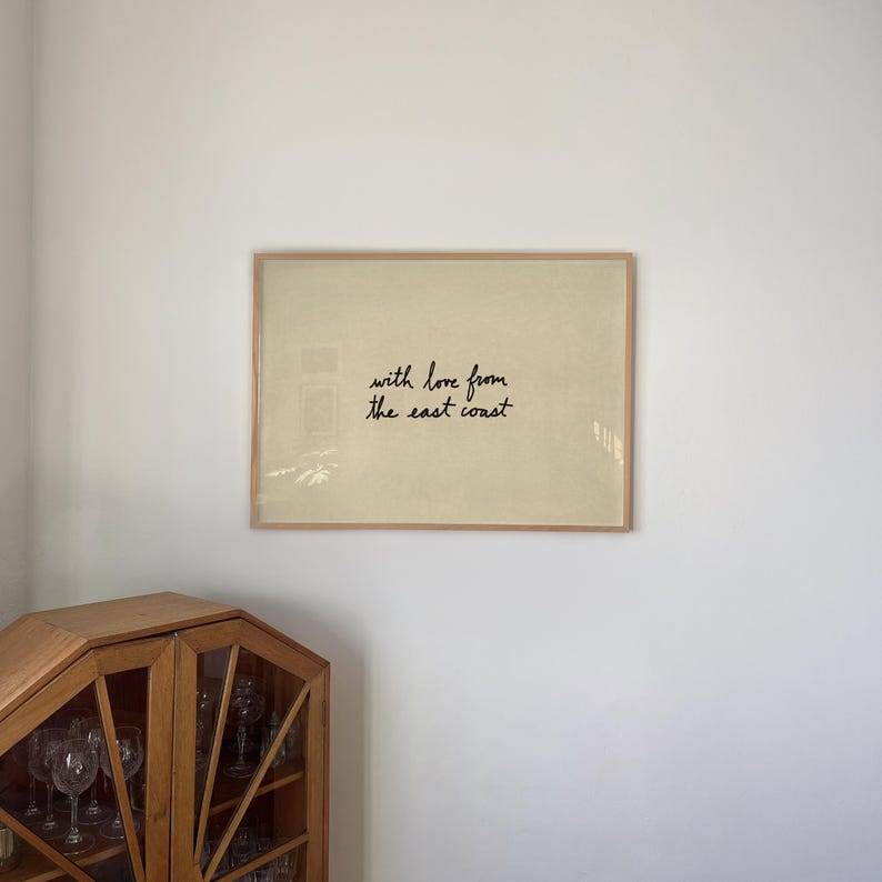 Puede incluir: Una impresi&oacute;n enmarcada con el texto "with love from the east coast" en escritura negra. La impresi&oacute;n tiene un fondo beige claro y un marco de madera natural. La impresi&oacute;n est&aacute; colgada en una pared blanca.