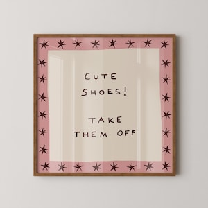 May include: A square framed wall art piece featuring a light beige centre with the dark brown text "CUTE SHOES! TAKE THEM OFF". The design is bordered by a pink frame decorated with a repeating pattern of dark brown hand-drawn star shapes.