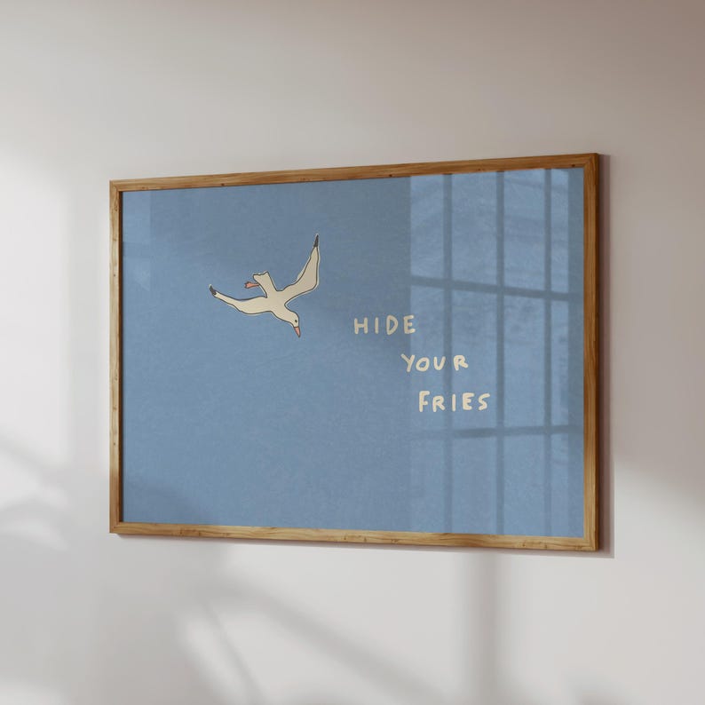 May include: A framed art print featuring a cartoon seagull in flight against a light blue background. The text "HIDE YOUR FRIES" is written in a playful font. The frame is made of light brown wood.