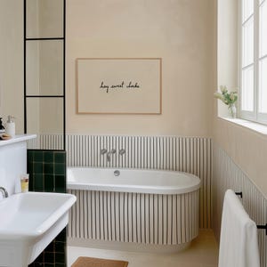 May include: A bathroom scene featuring a white oval bathtub with black vertical stripes on the wall. A framed print with the text "hey sweet cheeks" hangs above the tub. The room has a neutral color palette with a window and a sink.