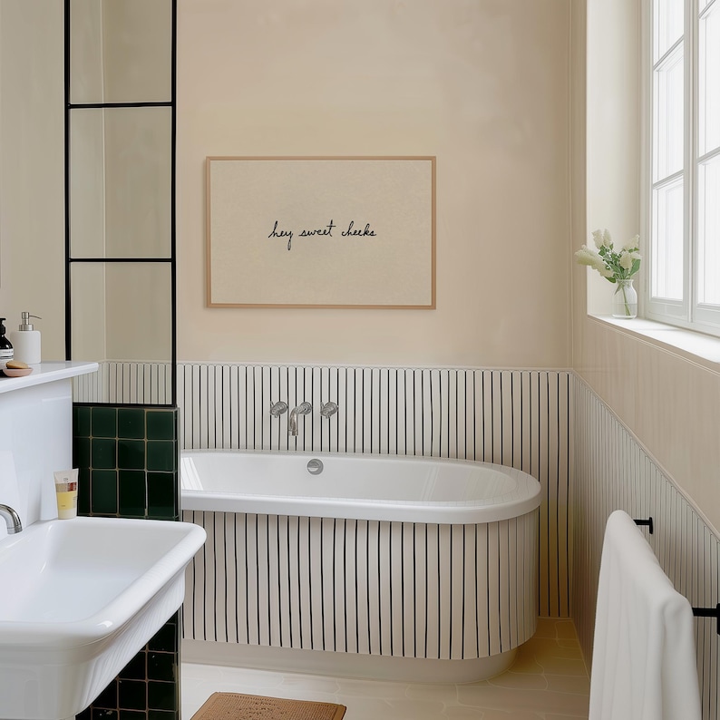 May include: A bathroom scene featuring a white oval bathtub with black vertical stripes on the wall. A framed print with the text "hey sweet cheeks" hangs above the tub. The room has a neutral color palette with a window and a sink.
