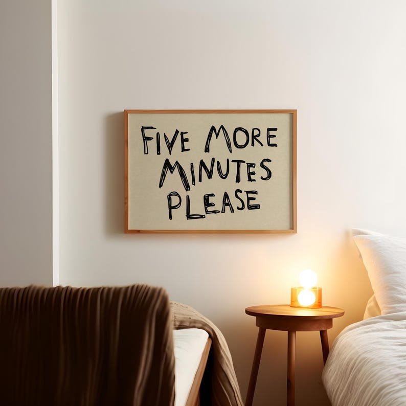 May include: A framed sign with the text "FIVE MORE MINUTES PLEASE" in black lettering. The sign is in a bedroom setting, with a bed and a small wooden side table with a lamp.