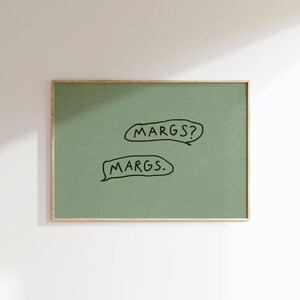 May include: A framed art print with a sage green background. The print features two speech bubbles with the text "MARGS?" and "MARGS." in black. The frame is a thin, gold color.