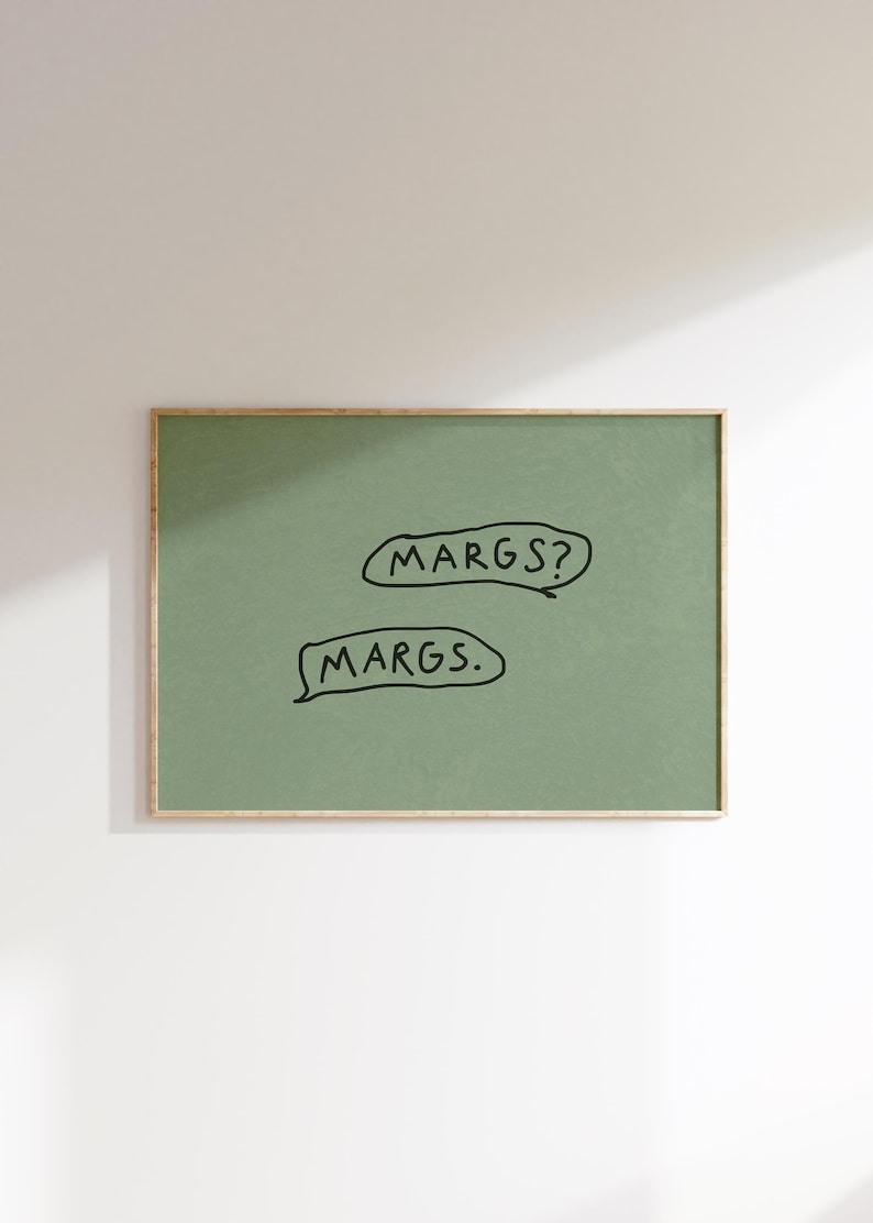 May include: A framed art print with a sage green background. The print features two speech bubbles with the text "MARGS?" and "MARGS." in black. The frame is a thin, gold color.