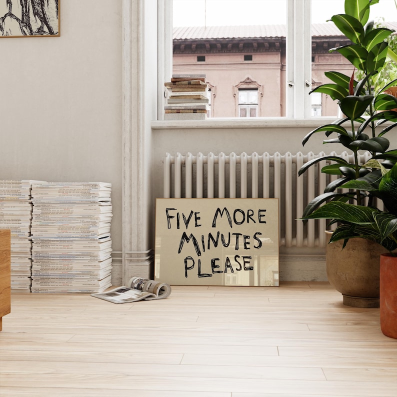 May include: A light-colored wooden floor with a sign that reads "FIVE MORE MINUTES PLEASE." A stack of magazines and a potted plant are also visible. The sign is leaning against a radiator.
