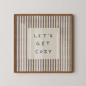 May include: A square wall art piece in a brown wooden frame featuring a beige center panel with the text "LET'S GET COZY" in black handwritten-style letters, surrounded by a border of vertical, textured, dark brown and beige stripes.