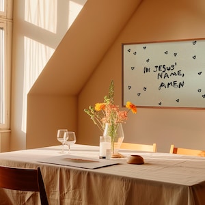 May include: A dining room scene with a wooden table set for a meal. A framed artwork on the wall reads "IN JESUS' NAME, AMEN" with heart doodles. A vase of flowers and two wine glasses are on the table.
