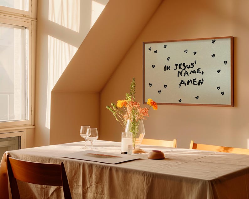 May include: A dining room scene with a wooden table set for a meal. A framed artwork on the wall reads "IN JESUS' NAME, AMEN" with heart doodles. A vase of flowers and two wine glasses are on the table.