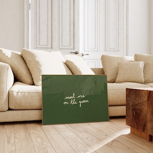 May include: A framed green artwork with the text "meet me on the green" in white script. The artwork is displayed on a wooden floor in front of a beige sofa and a wooden side table.