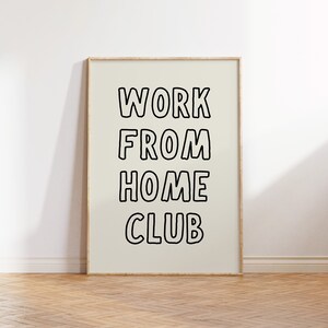 Op de afbeelding: Een ingelijste poster met de woorden "WORK FROM HOME CLUB" in vette, zwart omlijnde letters. De poster staat tegen een lichtbeige achtergrond en is omlijst in licht hout. De poster wordt weergegeven op een houten vloer.