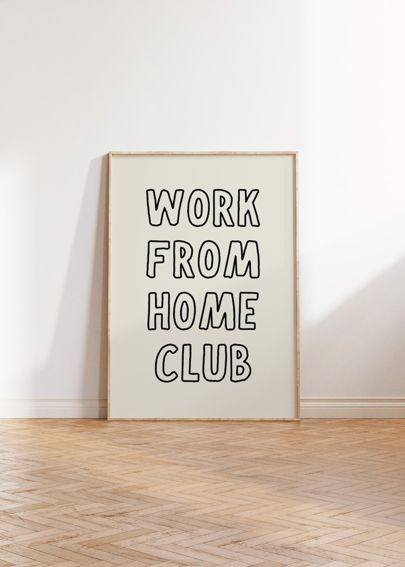 Op de afbeelding: Een ingelijste poster met de woorden "WORK FROM HOME CLUB" in vette, zwart omlijnde letters. De poster staat tegen een lichtbeige achtergrond en is omlijst in licht hout. De poster wordt weergegeven op een houten vloer.