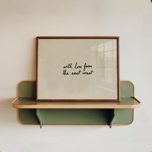 Puede incluir: Una impresi&oacute;n enmarcada con el texto "with love from the east coast" se encuentra en un estante verde y beige. La impresi&oacute;n tiene un marco marr&oacute;n y un fondo beige claro. El estante est&aacute; montado en una pared blanca.