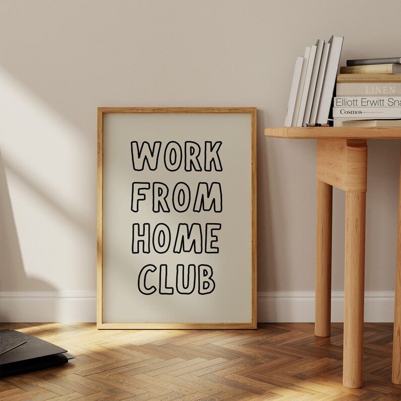 Op de afbeelding: Een ingelijste poster met de tekst "WORK FROM HOME CLUB" in zwarte, vette letters. De poster zit in een licht houten frame en leunt tegen een muur. Op de achtergrond is een houten tafel met boeken te zien en de vloer is van hout.