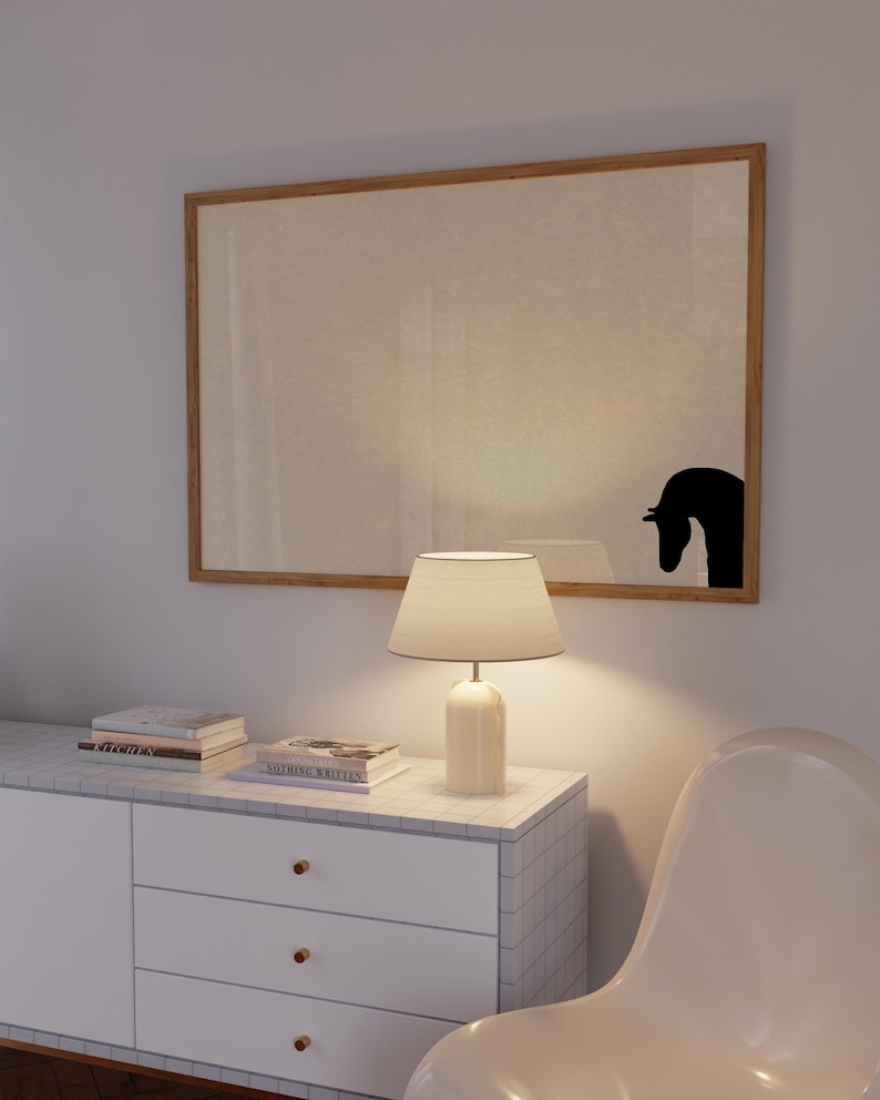 May include: A modern interior scene featuring a large framed artwork with a black horse silhouette. A white lamp with a beige shade sits on a white cabinet with gold knobs. Books are stacked on the cabinet. A white chair is in the foreground.