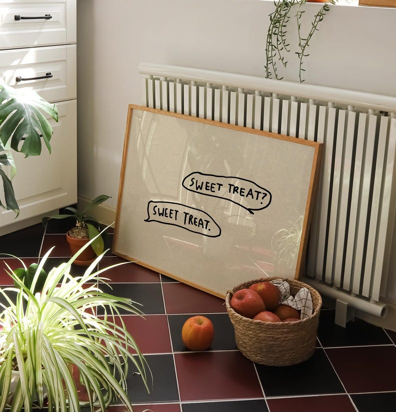 May include: A framed artwork with the text "SWEET TREAT?" and "SWEET TREAT." is propped against a white radiator. A woven basket filled with red apples sits nearby. A single apple rests on the floor. The scene is set on a red and black tiled floor.
