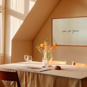 May include: A dining room scene with a wooden table set for a meal. A framed artwork with the text "vino, per favore" hangs on the wall. A vase of flowers and two wine glasses are on the table, which is covered with a beige tablecloth.