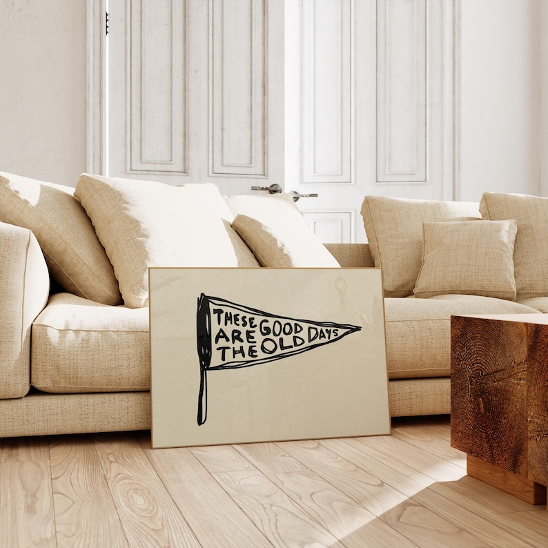 May include: A framed art piece with a beige background and black text that reads 'These are the good old days' in a pennant shape. The art is leaning against a beige couch with white pillows, and a wooden side table. The room has light wood flooring and white doors.