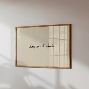 May include: A framed print with the phrase "hey sweet cheeks" in a handwritten style. The print has a light beige background and a natural wood frame. The image is hanging on a white wall.