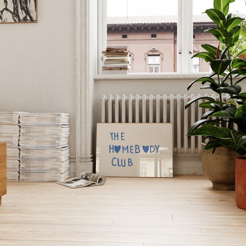 May include: A framed print with the text "THE HOMEBODY CLUB" in blue lettering with two blue hearts. The print is hanging on a white wall in a room with a wooden floor.