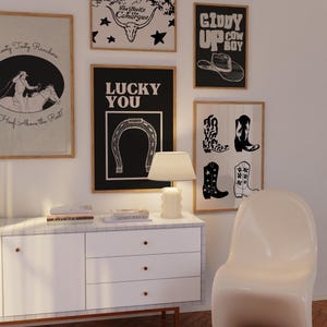 May include: A selection of framed art prints with a Western theme. The prints feature cowboy boots, a horseshoe, a cowboy hat, and text. The color scheme is mainly black, white, and tan. A white lamp and cabinet are in the foreground.