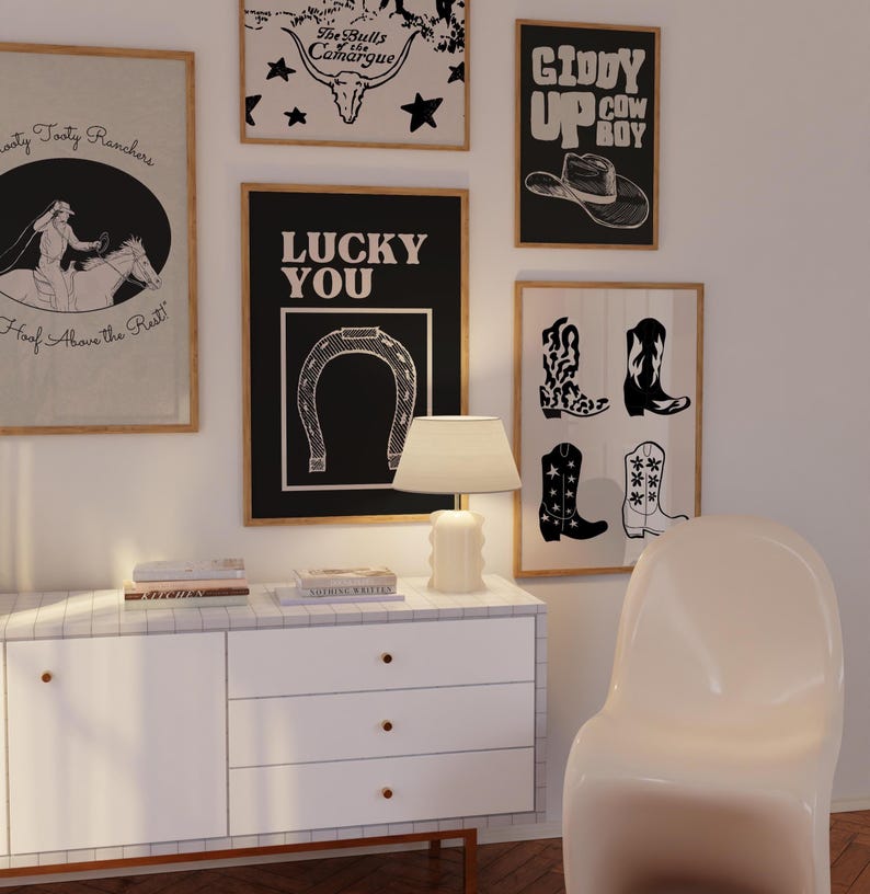 May include: A selection of framed art prints with a Western theme. The prints feature cowboy boots, a horseshoe, a cowboy hat, and text. The color scheme is mainly black, white, and tan. A white lamp and cabinet are in the foreground.