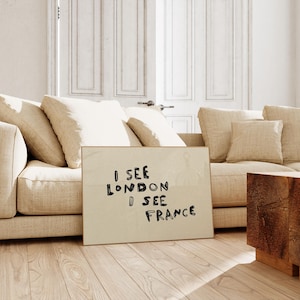 Puede incluir: Un cuadro enmarcado con el texto "I SEE LONDON I SEE FRANCE" en letras negras. El cuadro est&aacute; apoyado contra un sof&aacute; beige con varios cojines. Una mesa auxiliar de madera es visible a la derecha, y una puerta blanca est&aacute; en el fondo.