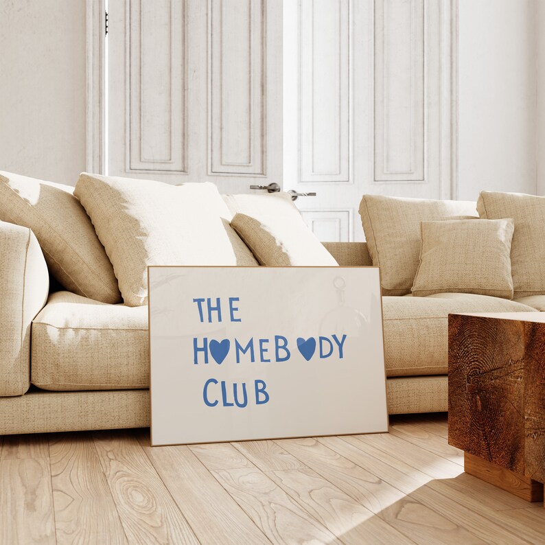 May include: A white sign with the text "THE HOMEBODY CLUB" in blue lettering with two blue hearts. The sign is leaning against a beige couch in a room with white walls and white doors.