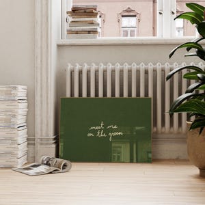 May include: A framed art print with a dark green background and the handwritten text "meet me on the green" in white. The print is displayed indoors, leaning against a wall near a window and a radiator.